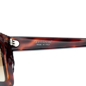 Op de afbeelding: Close-up van een bruine zonnebril met schildpadmotief. De tekst "Gianni Versace" en "MADE IN ITALY" zijn op de poot gedrukt. De bril heeft een zilveren scharnier.
