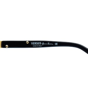 May include: Black Versus sunglasses arm with gold-colored accents. The text "VERSUS Gianni Versace" and "MADE IN ITALY" are printed on the arm. The sunglasses arm is curved and has a sleek design.