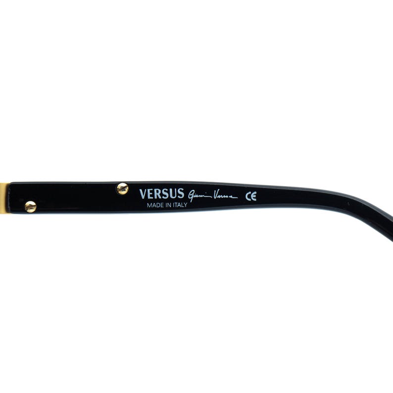 May include: Black Versus sunglasses arm with gold-colored accents. The text "VERSUS Gianni Versace" and "MADE IN ITALY" are printed on the arm. The sunglasses arm is curved and has a sleek design.