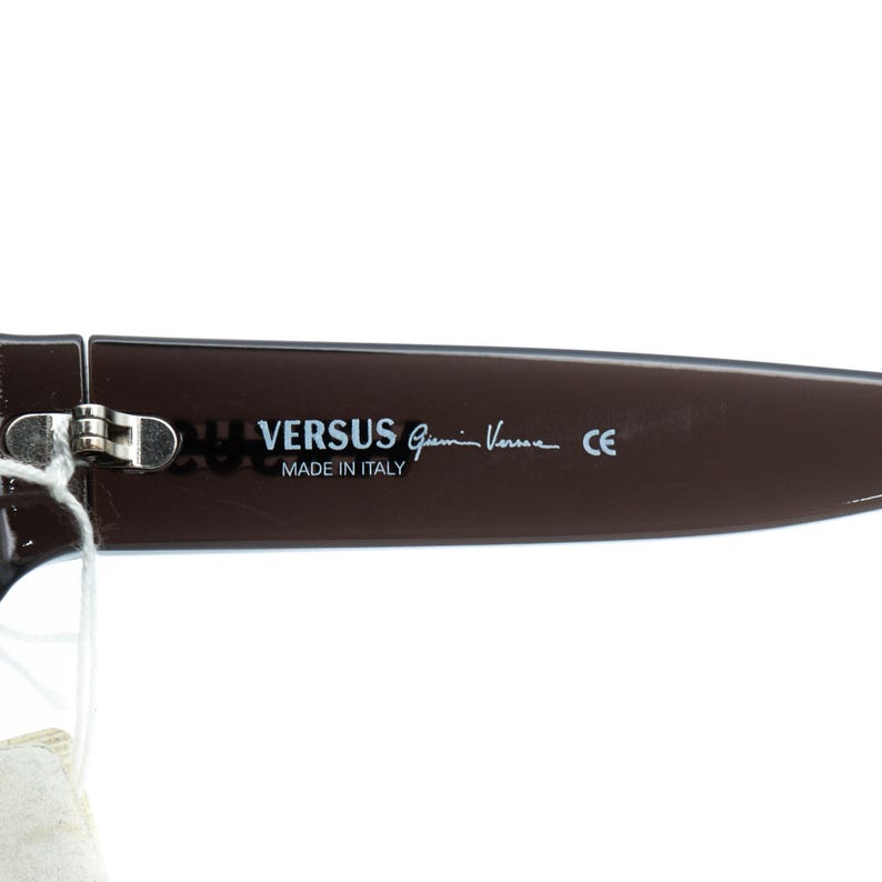 K&ouml;nnte beinhalten: Nahaufnahme eines braunen Versus Gianni Versace Sonnenbrillenb&uuml;gels. Der Text "VERSUS Gianni Versace" und "MADE IN ITALY" ist auf dem B&uuml;gel aufgedruckt. Der Sonnenbrillenb&uuml;gel hat eine dunkelbraune Farbe.