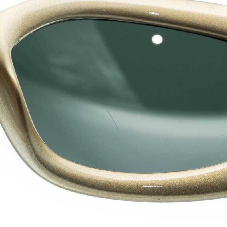 May include: Close-up of a pair of sunglasses. The frames are a light gold colour, with a rounded shape. The lenses are a dark, smoky grey. The sunglasses are set against a white background.