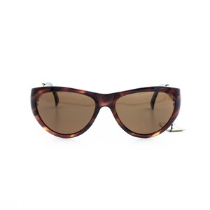 K&ouml;nnte beinhalten: Braune Sonnenbrille mit Schildpattmuster und braunen Gl&auml;sern. Die Sonnenbrille hat ein geschwungenes, umlaufendes Design. Der Rahmen hat ein gesprenkeltes Muster. Die Gl&auml;ser sind einfarbig braun. Die Sonnenbrille ist vor wei&szlig;em Hintergrund platziert.