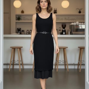 Puede incluir: Vestido negro sin mangas con corpiño ajustado y falda plisada. El vestido se ciñe a la cintura con un cinturón negro fino. La modelo lleva tacones negros.