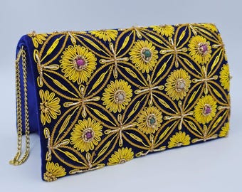 Puede incluir: Un bolso de mano rectangular de terciopelo azul real con un intrincado patrón de bordado de hilo amarillo en un motivo floral circular. El diseño está acentuado con pequeñas piedras de colores incrustadas e incluye una correa de cadena dorada.