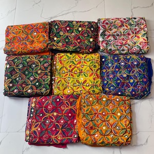 May include: A collection of colorful, embroidered textiles. Each piece features intricate patterns with mirrored accents. The textiles are folded, showcasing a variety of vibrant colors including orange, green, yellow, red, and blue. The overall aesthetic is decorative and ornate.