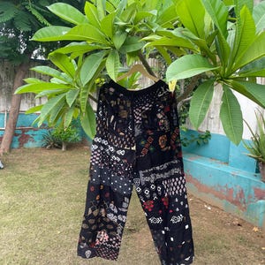 May include: Wide-leg trousers featuring a black base with a patchwork design of various white, red, and tan floral and geometric patterns. The pants have an elasticized waistband and a loose, flowing silhouette constructed from multiple fabric panels.