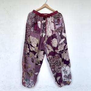 Könnte beinhalten: Patchwork-Hose mit einer Vielzahl von Blumen- und abstrakten Mustern in verschiedenen Violett-, Lavendel- und Weißtönen. Die Hose hat einen elastischen Bund mit Kordelzug und ist an einem Holzkleiderbügel aufgehängt.