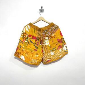 Könnte beinhalten: Senfgelbe Shorts mit Patchwork-Design, bestehend aus verschiedenen Blumen- und geometrischen Mustern in Rot-, Weiß- und Orangetönen. Die Shorts verfügen über einen elastischen Bund mit Kordelzug und eine lockere, entspannte Passform an den Beinen.