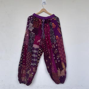 Könnte beinhalten: Weit geschnittene Hose mit Patchwork-Design in verschiedenen Violett-, Magenta- und Dunkelrot-Tönen. Die Hose hat einen elastischen Bund mit passendem violettem Kordelzug, und der Stoff zeigt zahlreiche komplexe florale und geometrische Muster.