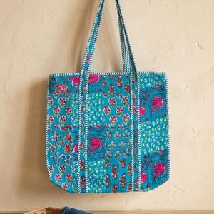 Könnte beinhalten: Eine türkisfarbene Tragetasche mit einem Blumenmuster in Pink, Rot und Blau. Die Tasche hat lange Träger und hängt an einem Ast. Das Design der Tasche umfasst eine Vielzahl von Blumen- und Paisley-Motiven.