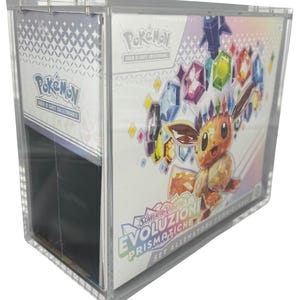 May include: A clear acrylic display case containing a Pokémon trading card game set. The set features a cartoon character and colourful gemstone graphics. Text includes "Evoluzioni Prismatiche" and "Set Allenatore Fuoriclasse".