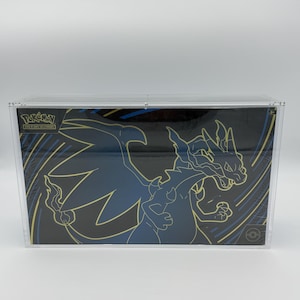 Peut inclure: Un coffret de cartes Pokémon avec un motif de dragon bleu et or sur fond noir, présenté dans un présentoir en plastique transparent. Le logo Pokémon est visible. Objet de collection.