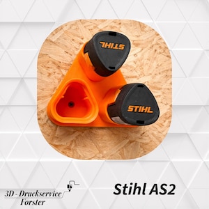Puede incluir: Un soporte de pared de plástico naranja para baterías Stihl AS2, con tres ranuras triangulares. Dos baterías negras Stihl AS2 están insertadas en el soporte, mientras que una ranura permanece vacía, revelando un orificio central para tornillo.