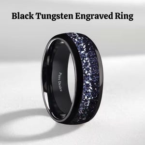 Könnte beinhalten: Schwarz gravierter Wolframring mit schwarzem Band und einem Mittelteil aus dunkelblauem und weiß gesprenkeltem Material. Der Ring trägt die Gravur "Pure Desire" an der Seite.