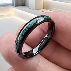 Könnte beinhalten: Ein schwarzer Ring mit einem Band aus schillerndem grün-blauem Inlay. Der Ring wird in einer Hand gehalten und trägt die Inschrift "Forever Yours" auf der Innenseite. Der Ring ist ein schlankes, modernes Design.