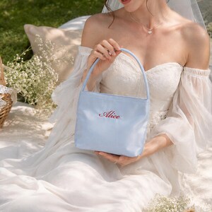 May include: A light blue handbag with a single top handle and the name Alice embroidered in red script on the front. The bag has a structured rectangular shape and a smooth finish, providing a simple and clean aesthetic for carrying small items.