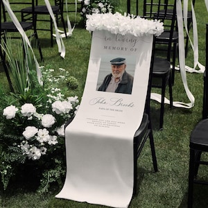May include: A white memorial chair sign draped over a black chair, featuring a photograph of a man, the text In Loving Memory Of John Brooks Papa of the Bride, and a quote. The sign is decorated with a cluster of white silk flowers at the top.