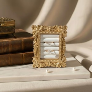 May include: Ornate gold-colored ring display frame with a white interior, holding two rings. The frame is on a white marble surface, with pearls scattered around. A small, decorative box and books are in the background.