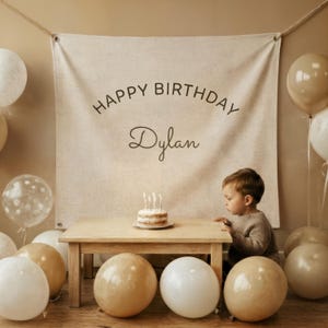 May include: A birthday celebration with a small cake and candles on a wooden table. A banner reading "HAPPY BIRTHDAY Dylan" is displayed. White and tan balloons are scattered around, with a child gazing at the cake.