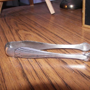 Op de afbeelding: Zilveren suikertang met een Art Deco-ontwerp. De tang is open, met de gebogen handvatten en het decoratieve patroon op de armen. De tang ligt op een houten oppervlak.