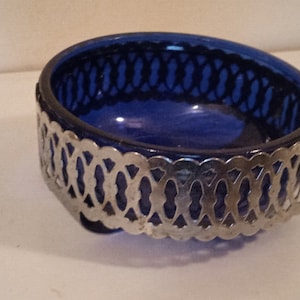 Op de afbeelding: Een kobaltblauwe glazen kom met een decoratieve zilverkleurige metalen filigraan overlay. De kom heeft een afgeronde vorm en staat op een wit oppervlak. Het metaalwerk heeft een open, herhalend patroon. Diameter: ongeveer 15 cm.