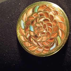 Può includere: Un oggetto decorativo rotondo con un bordo dorato e un motivo a spirale dettagliato nei toni del marrone, bianco e turchese. Il design ricorda un fiore o una spirale, con un bordo dorato metallico.