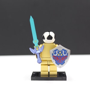 May include: A gold-colored toy figure with a soccer ball head, holding a blue sword and a shield with a red and blue design. The figure stands on a black base, against a black and white background.