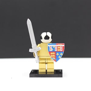 May include: A gold-colored toy figure with a soccer ball head, holding a silver sword and a shield with red, blue, and gold designs. The figure stands on a black base, showcasing a unique blend of sports and medieval themes.
