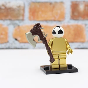 May include: A gold-colored Lego figure with a soccer ball head and a brown and silver axe. The figure stands on a black base against a brick background. The axe has a detailed handle and a silver blade.