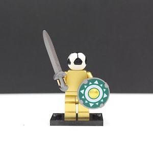 May include: A gold-colored toy figure with a soccer ball head, holding a silver sword and a green and white shield. The figure stands on a black base, with a black background.