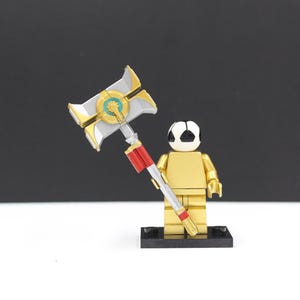 May include: A gold-colored minifigure with a black base holding a silver, gold, and red hammer. The minifigure has a white and black head. The hammer has a detailed design with a circular center.