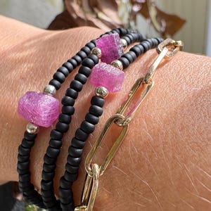 Peut inclure: Plusieurs bracelets superposés, composés de brins de perles noires entrecoupés de perles de pierres précieuses roses et d'accents argentés. Un bracelet à maillons dorés est également inclus. Les bracelets sont portés au poignet.