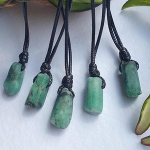 Puede incluir: Cinco colgantes de piedras preciosas verdes con cordones negros sobre un fondo blanco. Las piedras preciosas varían en forma, algunas naturales, otras pulidas. Los cordones están anudados en la parte superior.