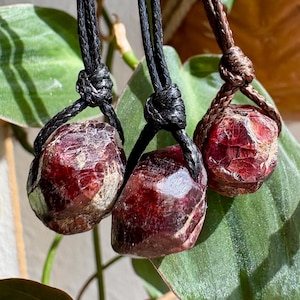 Peut inclure: Trois pendentifs en grenat brut rouge foncé suspendus à des colliers en cordon sombre. Chaque pierre présente une forme facettée et irrégulière avec des textures et des inclusions naturelles. Deux pendentifs sont sur des cordons noirs, un sur un cordon marron.