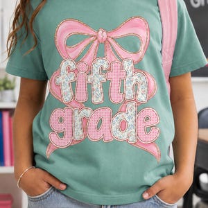 May include: A sage green t-shirt featuring a large pink bow graphic with gold glitter outlines. The text 'fifth grade' is printed in the center in a mix of blue floral and pink gingham patterns, with a pink backpack strap visible on the shoulder.
