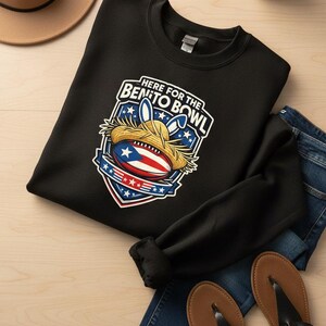 May include: Black crew neck sweatshirt with a graphic that reads "Here for the Benito Bowl." The graphic includes a football with the Puerto Rican flag, a straw hat, and bunny ears. The sweatshirt is shown with blue jeans and sandals.