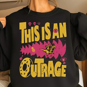 May include: Black sweatshirt with a yellow and pink graphic. The graphic includes the text "THIS IS AN OUTRAGE" in yellow, along with a cartoon dinosaur riding another dinosaur. Additional graphics include a pineapple, skulls, a bomb, and a trophy.