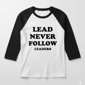 Może przedstawiać: Biała koszulka bejsbolowa z czarnymi rękawami. Koszulka zawiera napis "LEAD NEVER FOLLOW LEADERS" pogrubioną, czarną czcionką. Ma okrągły dekolt i rękawy o długości ¾.