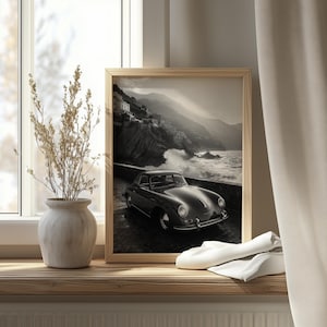 Pode incluir: Impressão emoldurada a preto e branco de um carro clássico numa estrada costeira. A imagem apresenta um veículo vintage com um fundo de montanhas e ondas. A impressão é exibida numa superfície de madeira com um vaso e tecido.