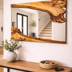 Op de afbeelding: Rechthoekige spiegel met een natuurlijke houten lijst. De lijst heeft een unieke, organische vorm met een lichtbruine kleur en donkerbruine accenten. De spiegel is gemonteerd op een witte muur boven een houten tafel.