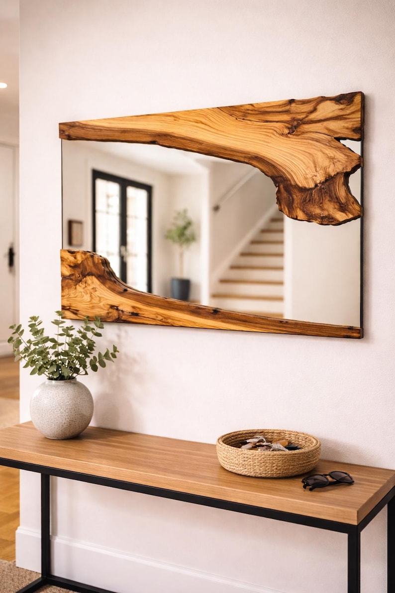 Op de afbeelding: Rechthoekige spiegel met een natuurlijke houten lijst. De lijst heeft een unieke, organische vorm met een lichtbruine kleur en donkerbruine accenten. De spiegel is gemonteerd op een witte muur boven een houten tafel.