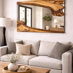 Op de afbeelding: Een rechthoekige spiegel met een houten lijst met een levende rand. De lijst is lichtbruin van kleur met een natuurlijk houtnerfpatroon. De spiegel hangt boven een lichtgekleurde bank met decoratieve kussens.