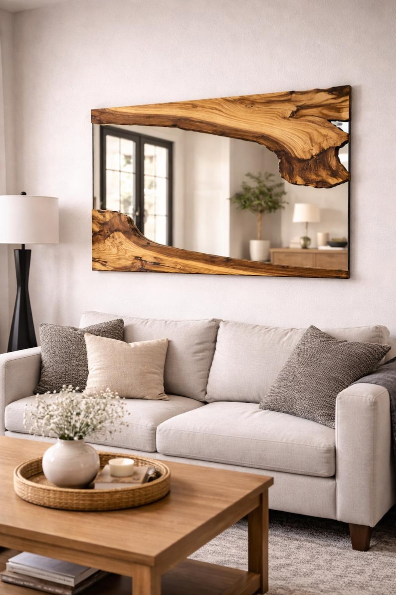 Op de afbeelding: Een rechthoekige spiegel met een houten lijst met een levende rand. De lijst is lichtbruin van kleur met een natuurlijk houtnerfpatroon. De spiegel hangt boven een lichtgekleurde bank met decoratieve kussens.