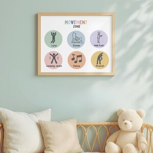 May include: A framed print titled "MOVEMENT ZONE" with colorful circles illustrating various activities: Jump, Stomp, Wall Push, Jumping Jacks, Dance, and Stretch. The print is in a light wood frame, hanging on a light blue wall above pillows and a teddy bear.