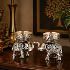 Peut inclure: Deux bougeoirs argentés en forme d'éléphant avec des détails ornés. Chaque éléphant soutient un bougeoir en forme de bol. Les éléphants se font face, leurs trompes se touchant. Un Bouddha et une bougie allumée sont en arrière-plan.