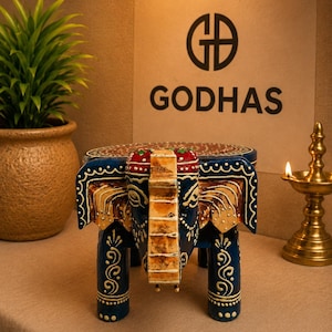 Puede incluir: Un pequeño taburete de madera pintado a mano con forma de elefante. El taburete presenta un cuerpo azul profundo con detalles pintados en blanco y oro, un tocado rojo y dorado, y una parte superior circular con un diseño. El texto GODHAS es visible.