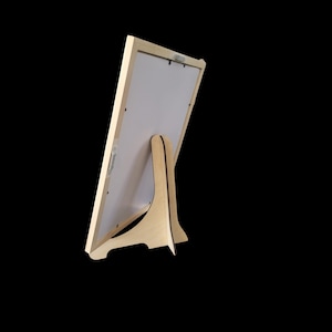 May include: A rectangular picture frame made of light-colored wood, shown from the back. The frame features a white backing board and a sturdy, integrated wooden stand attached to the center, allowing the frame to be displayed in an upright position.