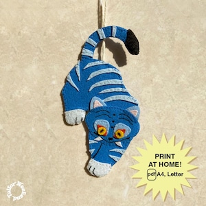 May include: A blue felt cat ornament with white stripes and accents, hanging from a string. The cat has yellow eyes and a black tail tip. A yellow starburst graphic with the text "PRINT AT HOME! pdf A4, Letter" is in the lower right.