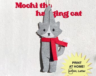May include: A gray felt cat ornament with a red scarf, hanging from a string. The cat has white markings and a black nose. Text above reads "Mochi the hanging cat." A yellow burst graphic says "PRINT AT HOME! pdf A4, Letter."