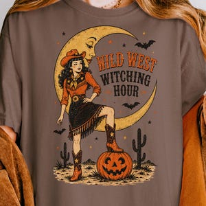 May include: An espresso-colored t-shirt featuring a graphic of a cowgirl standing with one foot on a jack-o'-lantern. The design includes a crescent moon, bats, cacti, and the text "WILD WEST WITCHING HOUR" in a stylized orange and black font.